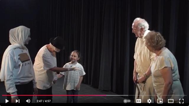 The image is a still from a video showing a scene on a stage. There are five people: two adults in white, appearing to be mime artists (one wearing a headscarf, the other a hat), a young girl in a light-colored shirt, and an elderly couple, the man slightly hunched and the woman beside him. The scene suggests a performance or play, possibly dealing with themes of age, youth, and perhaps the passing of time or generational differences. The dark backdrop emphasizes the figures in the foreground.