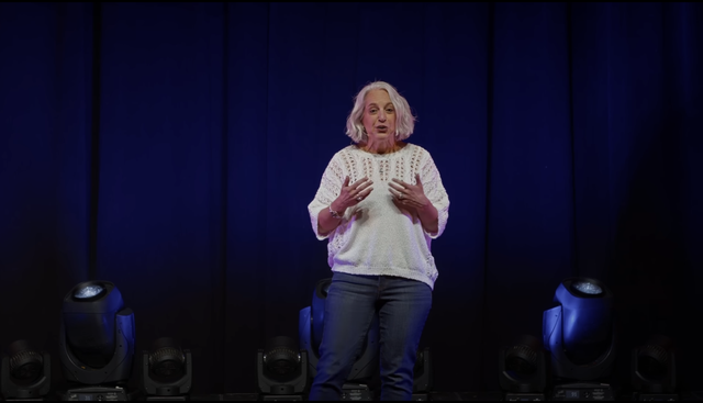 A woman with short, gray hair is standing in front of a dark blue curtain. She is wearing a white crochet sweater and blue jeans. She appears to be speaking, and her hands are gesturing. There are several large stage lights on the stage floor in front of her. The image is likely a still from a video. 