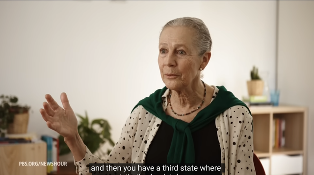 An older woman with gray hair, wearing a white shirt with black polka dots and a green shawl, gestures with her right hand as she speaks. She is seated in a room with a bookshelf in the background. The room is brightly lit and the woman is looking towards the camera. The caption "and then you have a third state where" appears at the bottom of the image.  The overall tone of the image is informative and engaging.