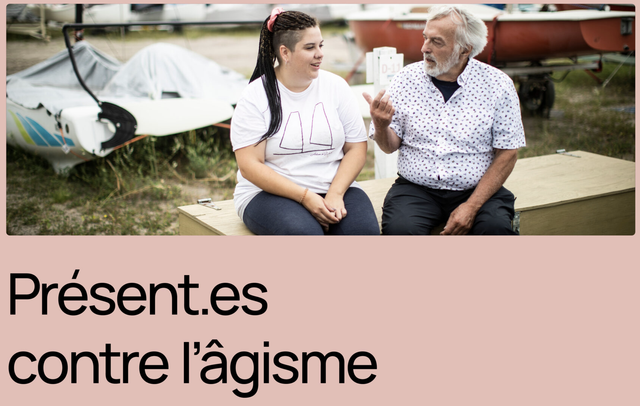 A young woman with braids and a pink headband sits on a wooden box, listening intently to an older man with a white beard and a patterned shirt. They are both smiling, suggesting a relaxed and comfortable conversation. The woman is wearing a white t-shirt with a simple design and black pants, while the man is wearing a white shirt with a patterned design and black pants. The background features a blurred image of a boat and a grassy area, creating a sense of outdoor setting. The image is accompanied by the text "Présent.es contre l'âgisme", which suggests a message of solidarity and resistance against ageism. The overall atmosphere of the photo is warm and positive, highlighting the importance of intergenerational connection and respect.