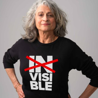 A woman with flowing grey hair wearing a black shirt that has the word invisible in white lettering on it stacked into three parts, "in", "vis", and "able" with the word "in" crossed out by a large red X. The look on her face is both defiant and satisfied.