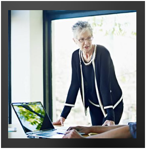 A feminine presenting person leaning over a desk and computer. They are speaking to someone sitting nearby.