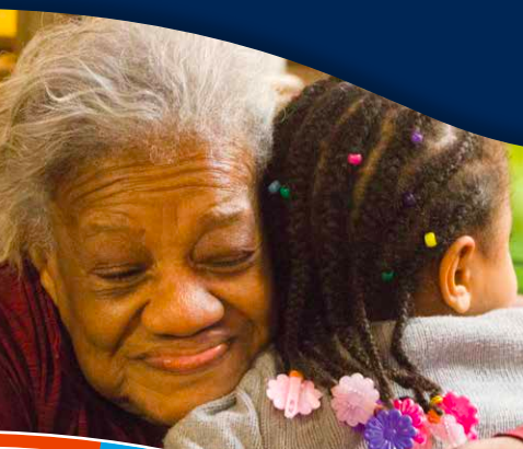 A woman with grey hair smiling while hugging a younger person who has braids in their hair with flowers on the ends of the braids.