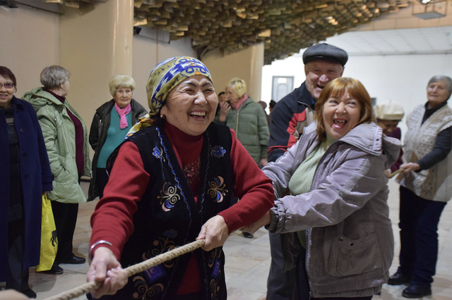 Eye-level view of a group of mostly older adults engaged in a tug-of-war. The central focus is two women intensely pulling on a thick rope. One woman is wearing a vibrant red sweater and a traditional-looking, dark vest with embroidery; the other is wearing a light purple jacket. The group includes a diverse range of people, mostly women, of different ethnicities and appearances. Some are wearing casual clothing, while others have more traditional or cultural attire. There's a man in a newsboy cap participating as well, and another man in a darker jacket.