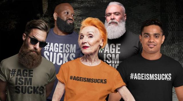A group of people wearing shirts that have the hashtag "ageism sucks" on them. In the center is a woman with red hair and an orange shirt. Behind her are two men wearing black and grey shirts. The man wearing the black shirt has a long grey beard, while the man wearing the grey shirt has a short black beard.