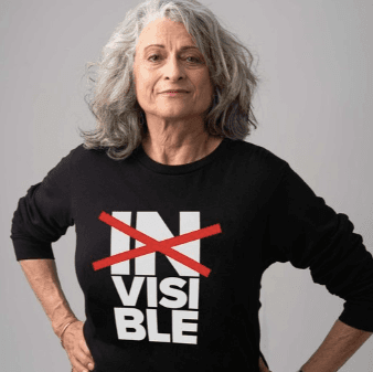 A woman with flowing grey hair wearing a black shirt that has the word invisible in white lettering on it stacked into three parts, "in", "vis", and "able" with the word "in" crossed out by a large red X. The look on her face is both defiant and satisfied.