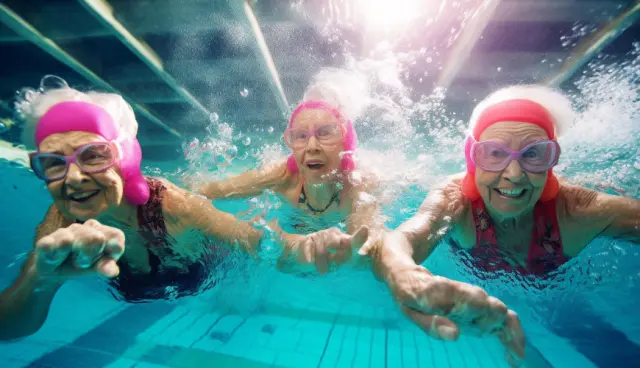 Three women swimming under water in a large swimming pool. All three are wearing pink swimming caps.