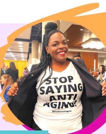 A smiling woman wearing a jacket that she is holding open to show a white shirt with black letters that says "Stop saying anti aging!" and under that "@thedementiaguru"
